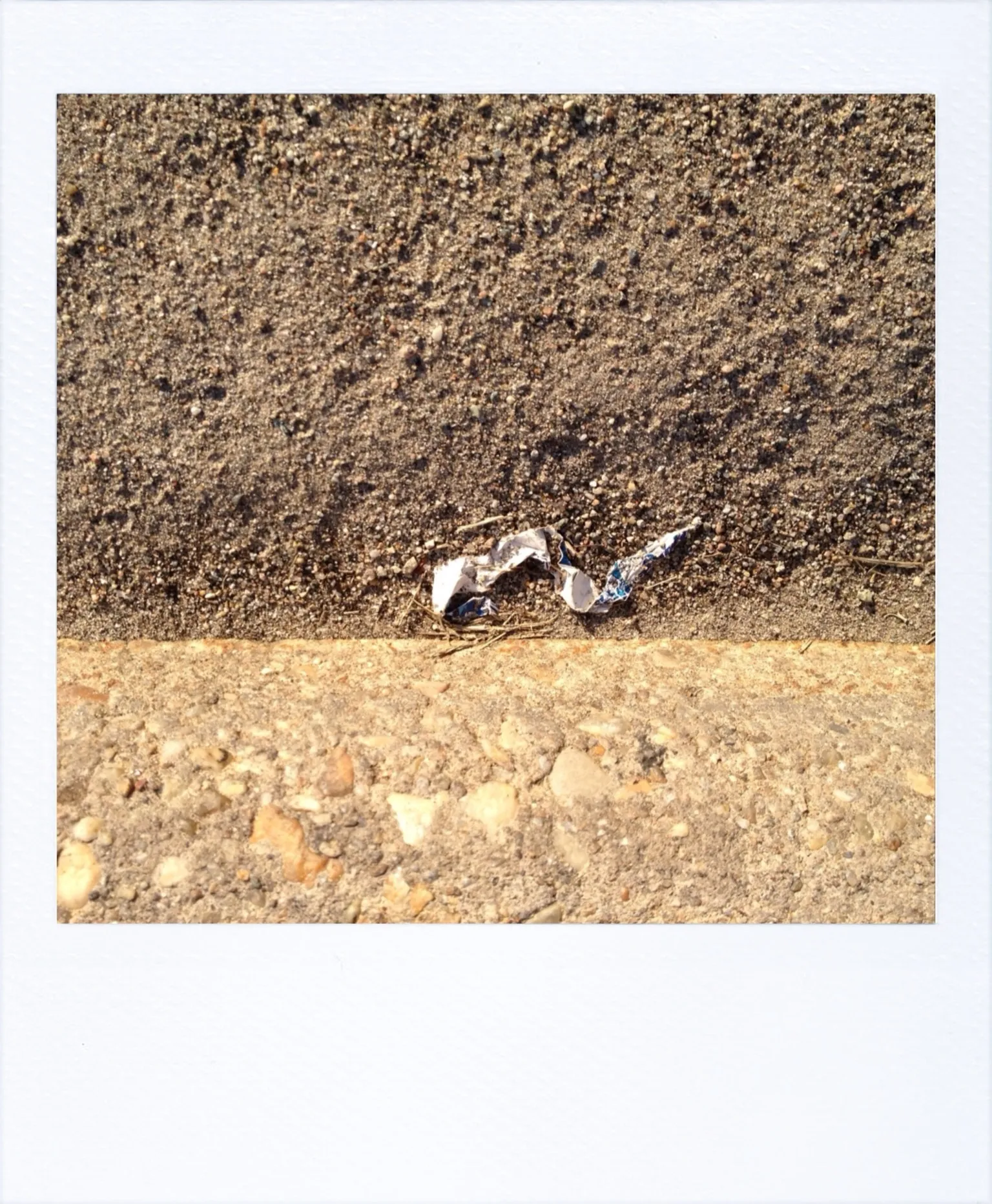 A small piece of crumpled foil lies in the seam between two rough concrete surfaces in bright sunlight.