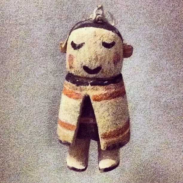 Small handcrafted figure with a painted face, closed eyes, and a striped robe, hanging against a textured background.