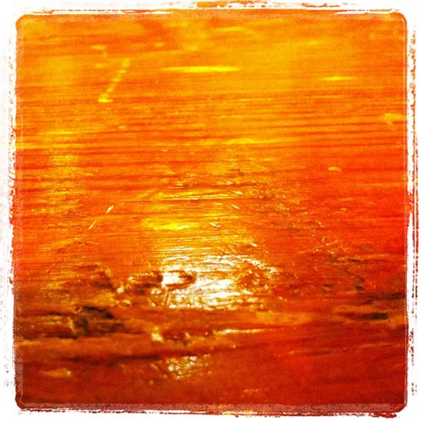 Close-up of a warm-toned wooden surface with visible grain, scratches, and glossy reflections under lighting.