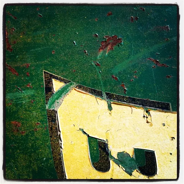 Close-up of a weathered green surface with peeling paint and a partially obscured yellow and black letter or symbol.
