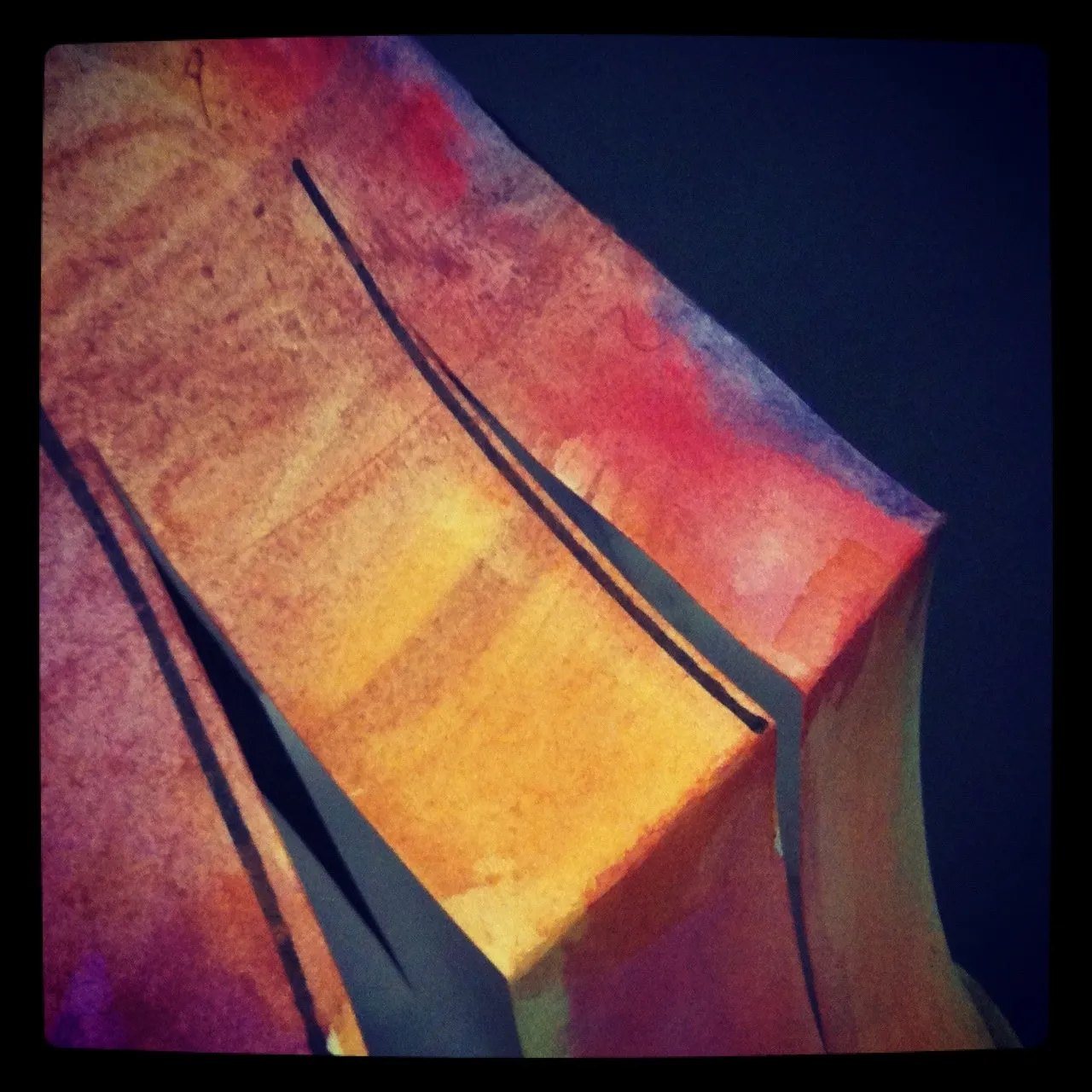Close-up of a painted, folded paper sculpture in warm tones of orange, red, and purple against a dark background.