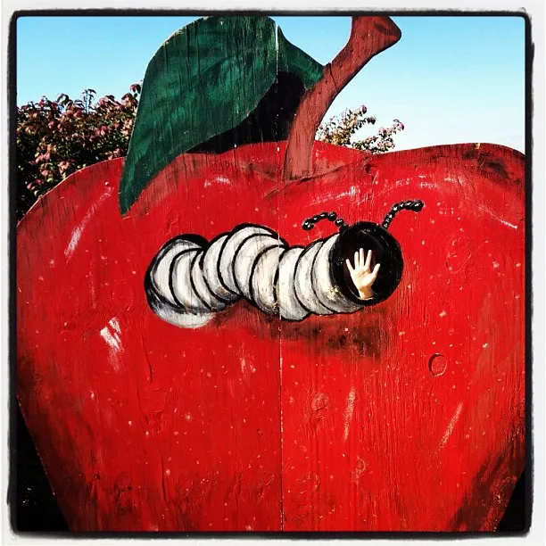 Painted wooden cutout of a red apple with a cartoon worm, featuring a hole where a child’s hand emerges playfully.