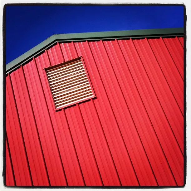 Close-up of a red metal barn wall with vertical ridges and a square vent, set against a deep blue sky.