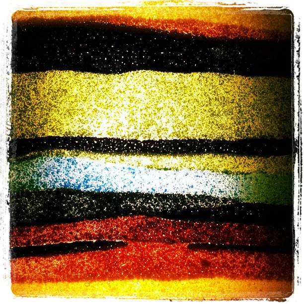 Close-up of layered colored sand in horizontal bands of red, yellow, green, blue, and black, creating a textured, abstract pattern.