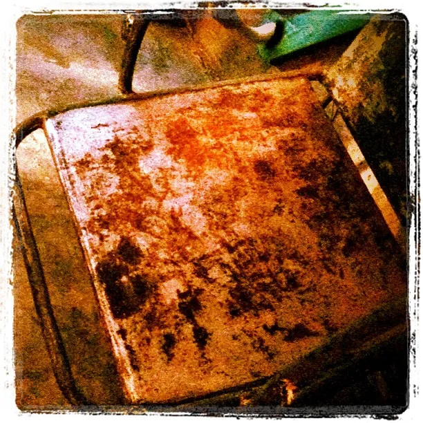 Close-up of a heavily rusted metal chair seat with worn paint, set against a concrete floor in a dimly lit space.