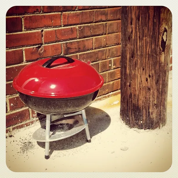 Small red and black charcoal grill with lid closed, placed on a sidewalk next to a brick wall and wooden utility pole.