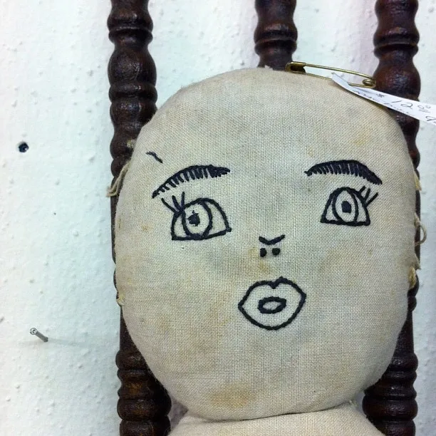A handmade cloth doll with exaggerated facial features drawn in black ink, including wide eyes, arched brows, and pursed lips.