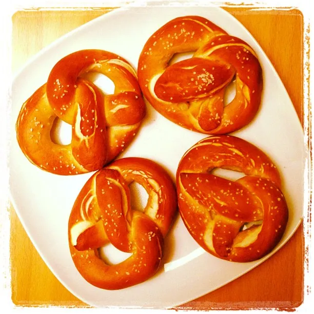 Four golden-brown soft pretzels with coarse salt, arranged on a white triangular plate on a wooden table.