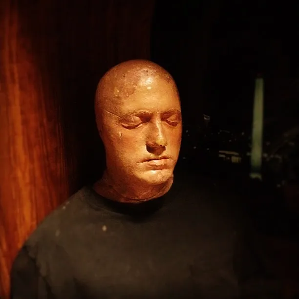 Wax or plaster bust of a bald man with eyes closed, wearing a dark shirt and lit dramatically from above.