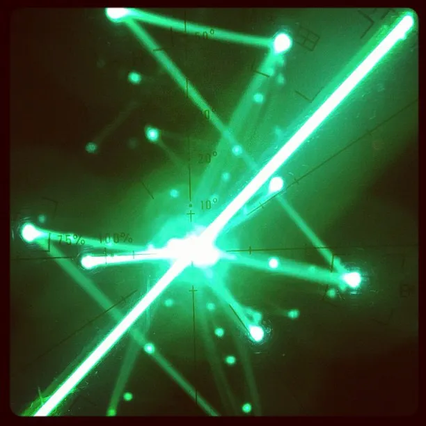 Bright green laser beams intersect at various angles on a grid-marked surface, creating a glowing starburst pattern.