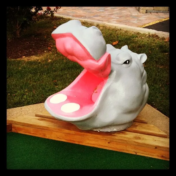 Large gray and pink hippopotamus head sculpture with open mouth, part of a miniature golf obstacle on green turf.