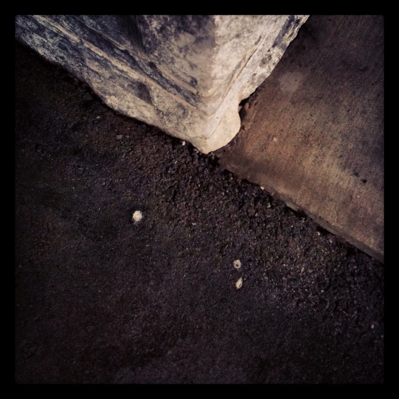 Close-up of the base of a stone column at the intersection of rough asphalt and smooth concrete pavement, with scattered debris.