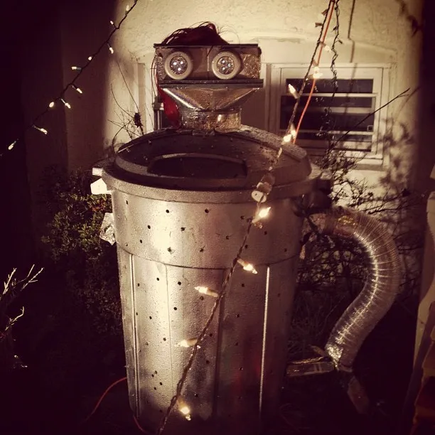 Homemade robot costume or sculpture made from a trash can and ducting, decorated with lights and placed outside a house.