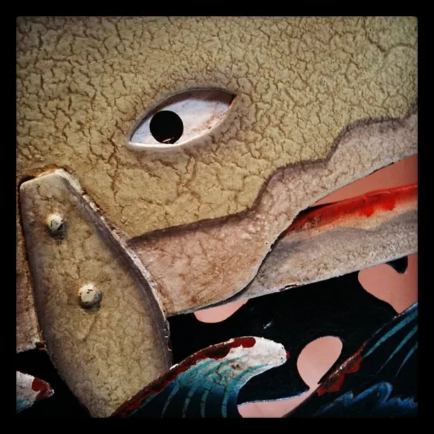 A close-up of a weathered and painted wooden surface featuring a stylized eye and mouth, likely part of a decorative fish or sea creature design.