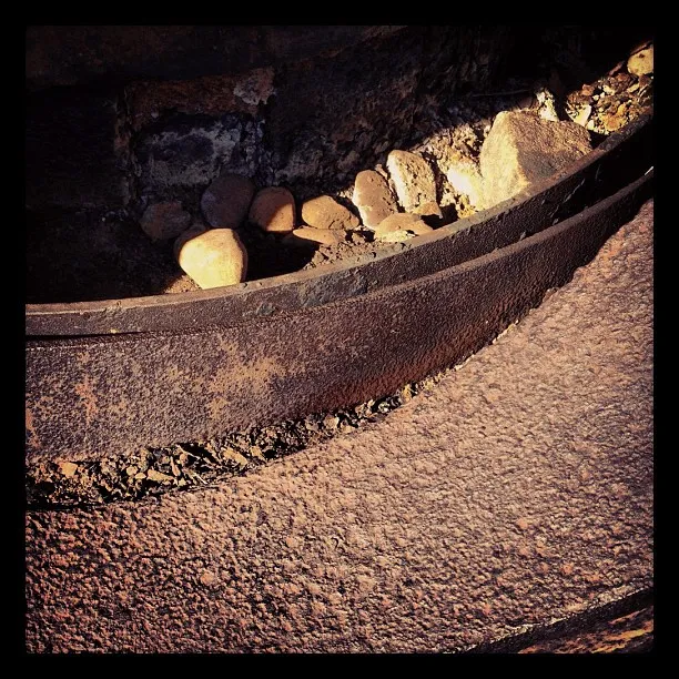 Close-up view of a weathered metal surface curving around a shallow stone pit, with scattered rocks and textured rust.