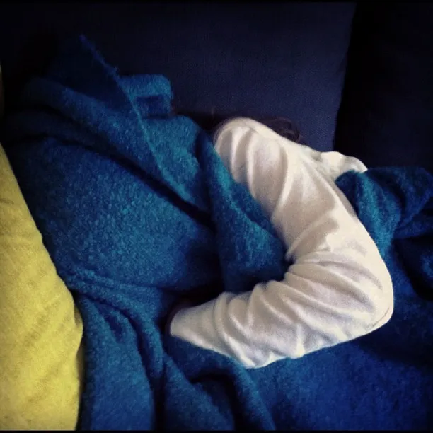 Child lying on a couch, mostly covered by a textured blue blanket, with only an arm and part of the head visible.