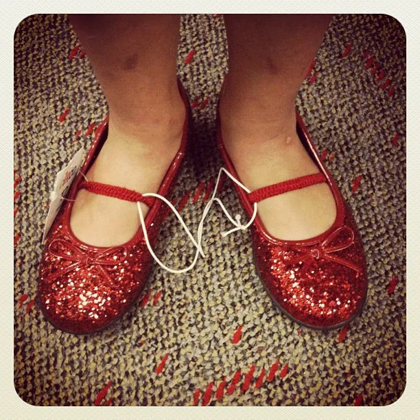 Child’s feet wearing sparkly red Mary Jane shoes with price tags and security ties on a speckled carpeted floor.
