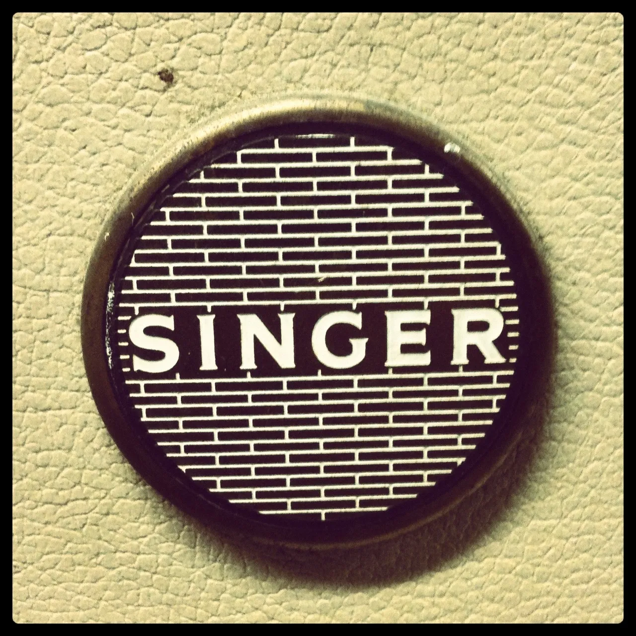 Close-up of a circular metal badge with the word “SINGER” in bold white letters over a black background with horizontal white lines.