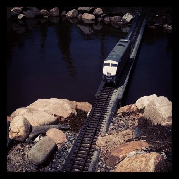 A model train crosses a narrow bridge over dark water, surrounded by scattered rocks and gravel on the riverbank.