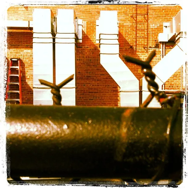 Industrial rooftop scene with metal air ducts mounted on a red brick wall, viewed through a foreground of barbed wire and black pipe.