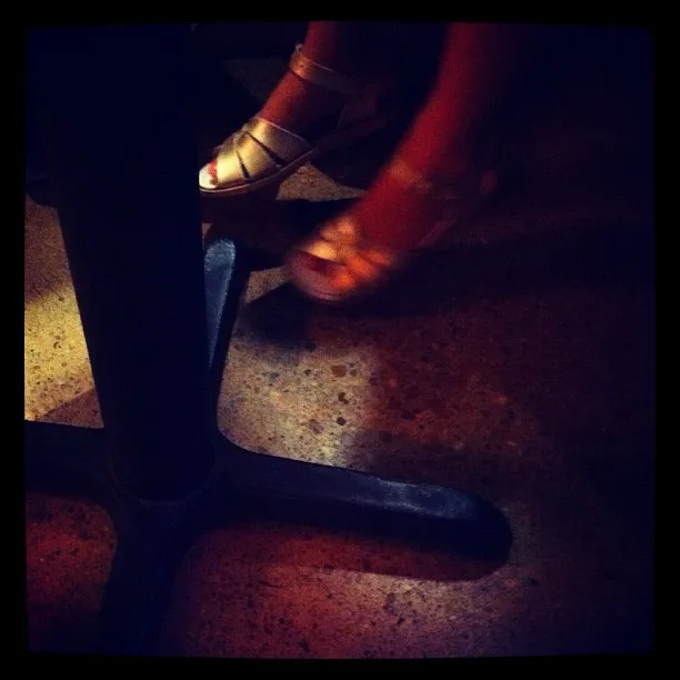 Dimly lit scene showing a pair of feet in gold sandals beside a metal table base on a textured concrete floor.