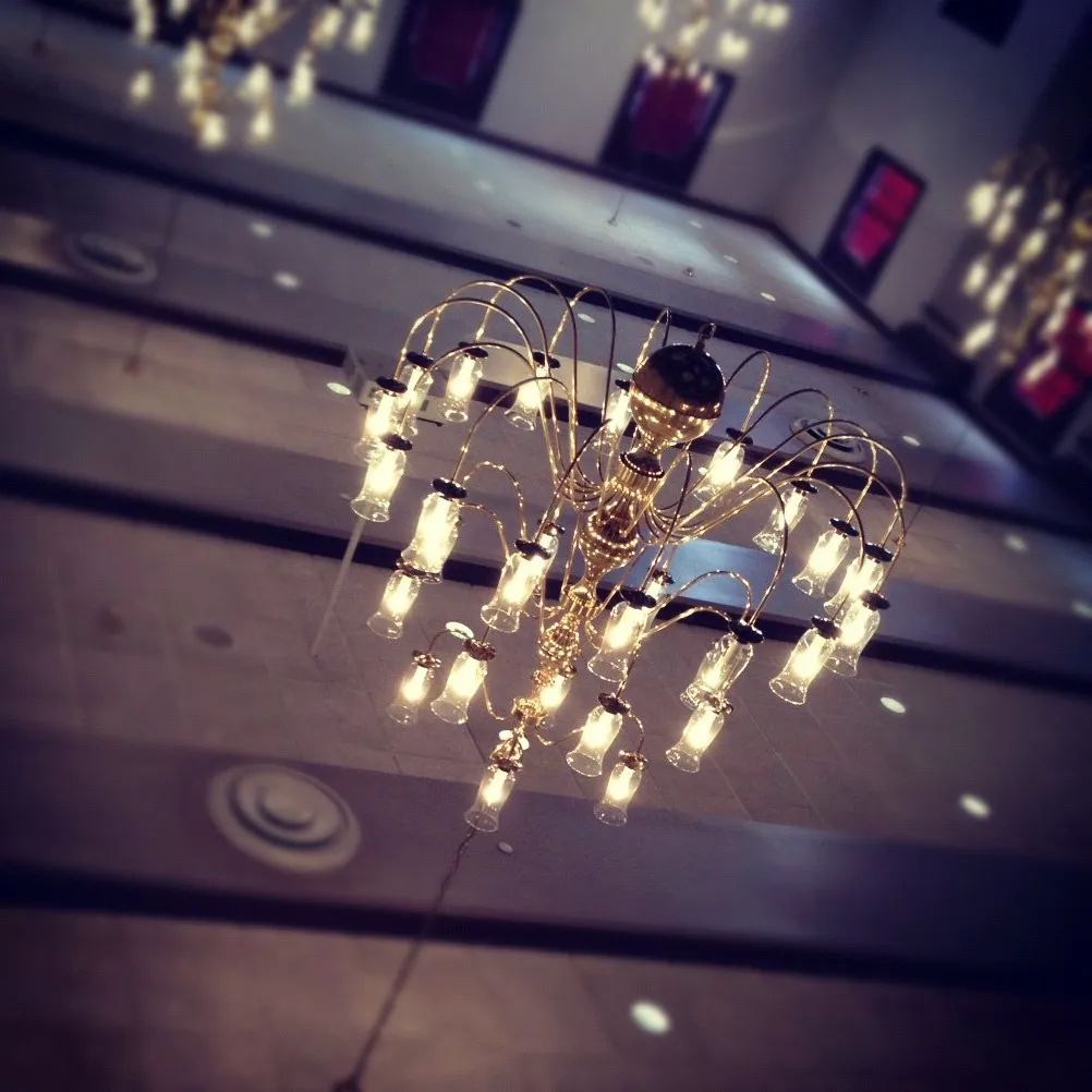 Large gold chandelier with multiple lit bulbs hanging from a high ceiling, reflected in a polished surface below.