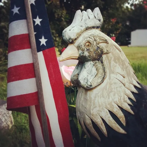 Close-up of a carved wooden rooster next to a small American flag in an outdoor setting with grass and flowers.