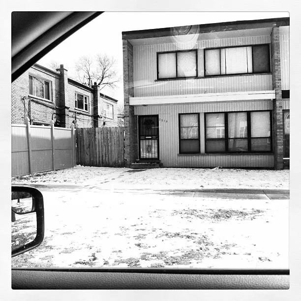 Black and white photo of a two-story building with closed blinds, viewed from inside a car across a snowy street.