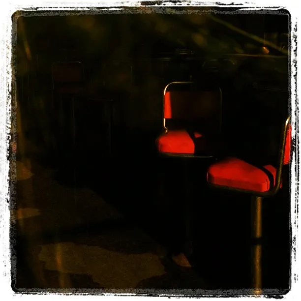 Dimly lit scene with red diner-style chairs partially illuminated by a warm light, surrounded by deep shadows.