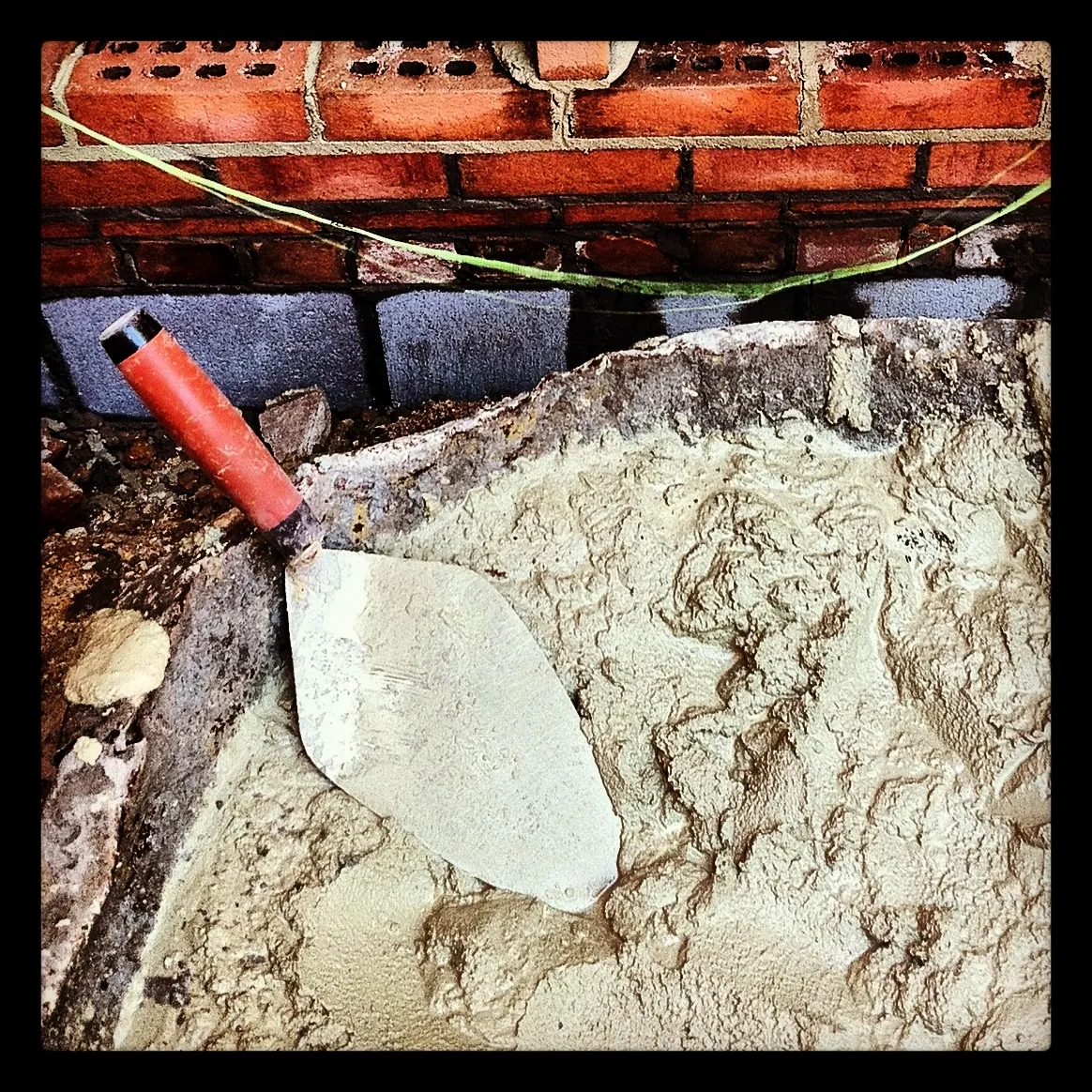 Close-up of a red-handled trowel resting in a pile of mixed cement in front of a partially built brick wall with visible mortar lines.