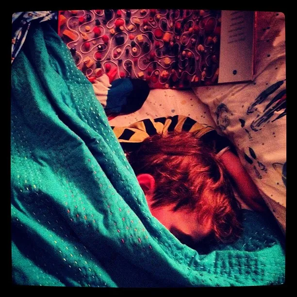 Child with reddish-brown hair sleeping under a teal blanket, surrounded by patterned bedding and an open book.