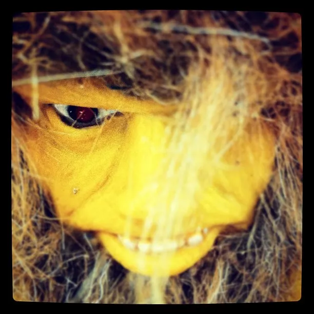 Close-up of a yellow-faced doll or mask with artificial hair, red eyes, and an unsettling grin, partially obscured by strands of hair.
