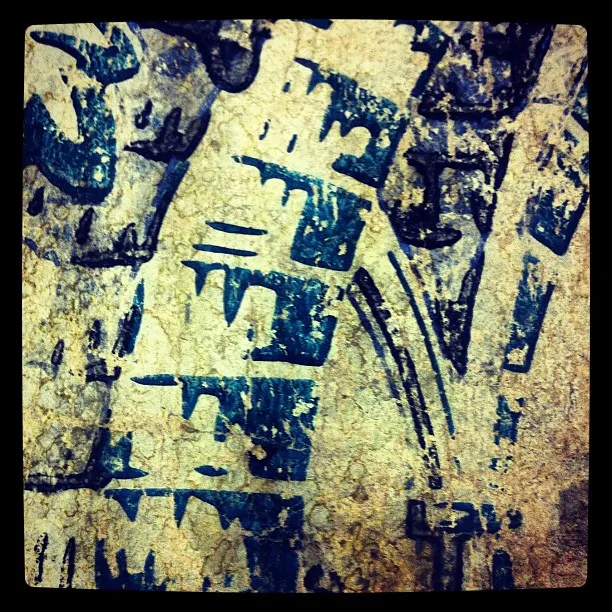 Close-up of weathered, cracked surface with faded blue geometric print or symbols on tan stone or plaster background.