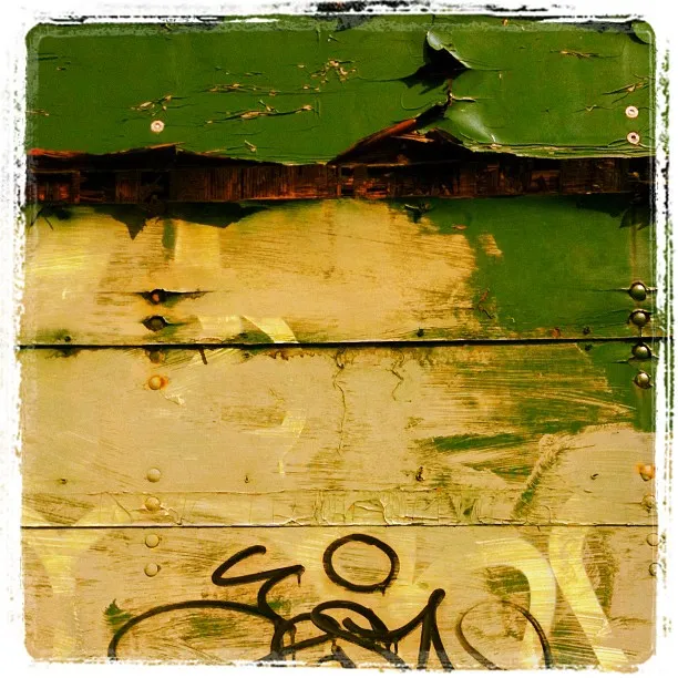 Close-up of weathered green and beige wooden surface with peeling paint, rusted nails, and a black bicycle graffiti symbol.