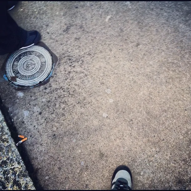 Two shoes visible on a sidewalk near a city water access cover, with a cigarette butt on the curb in the corner.