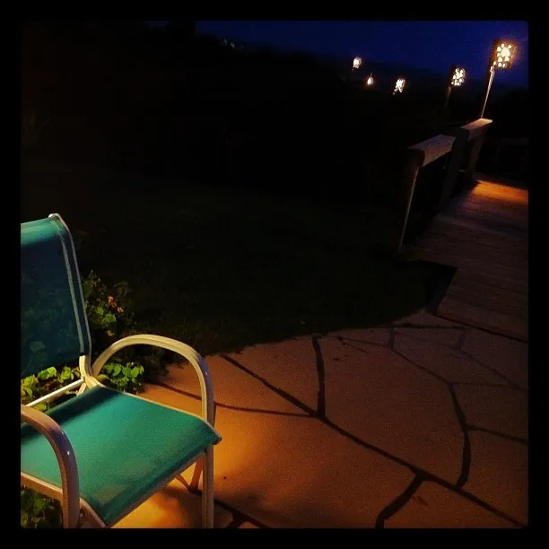 A turquoise metal chair on a stone patio at night, with a wooden walkway lit by decorative lights in the background.