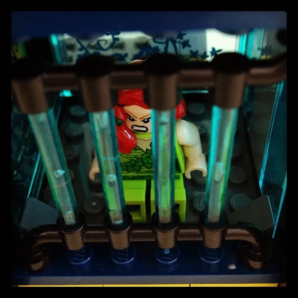 LEGO minifigure of Poison Ivy with red hair and a green outfit, posed behind translucent jail bars in a toy prison cell.