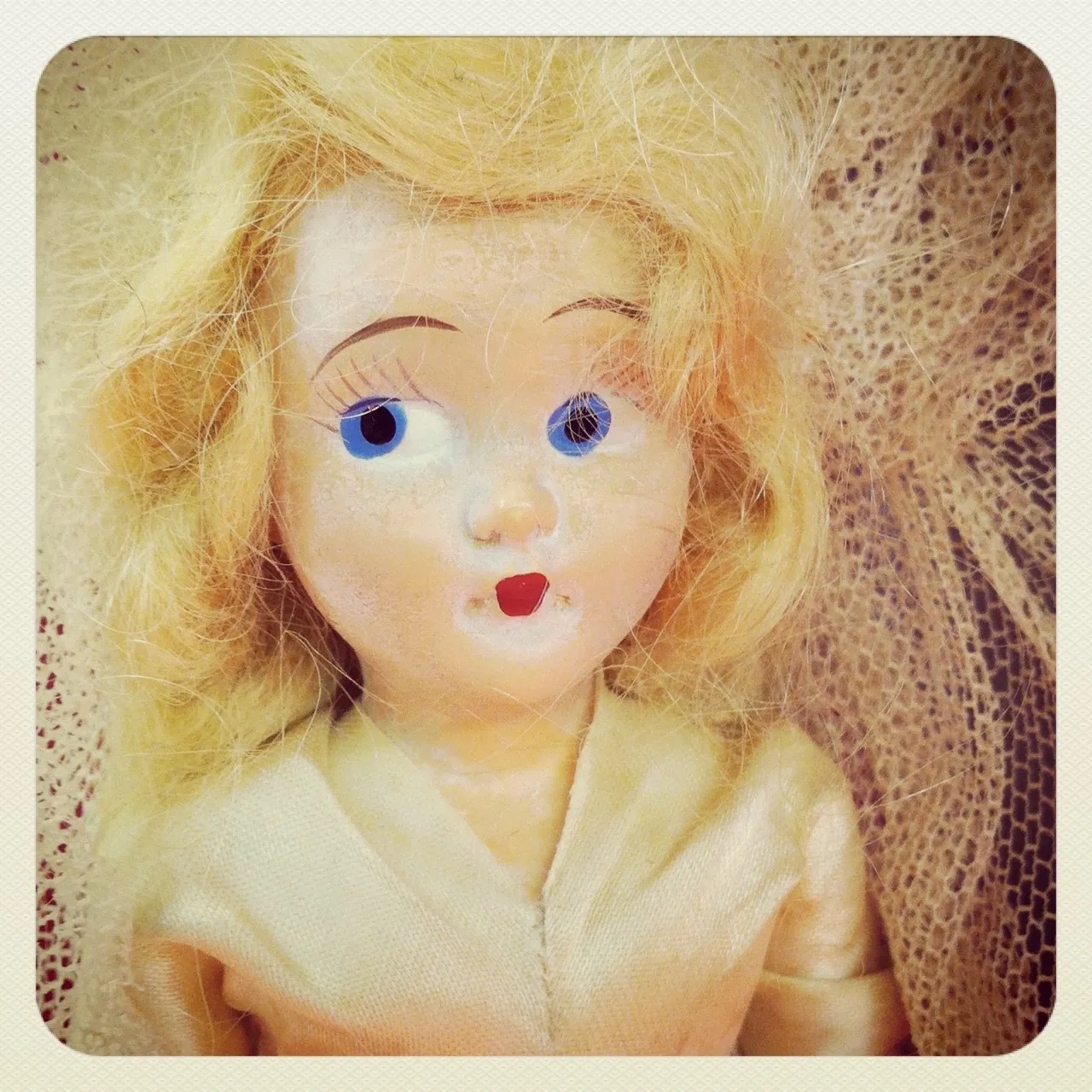 Close-up of a vintage blonde doll with wide blue eyes, red lips, and a cream-colored dress, posed against lace fabric.