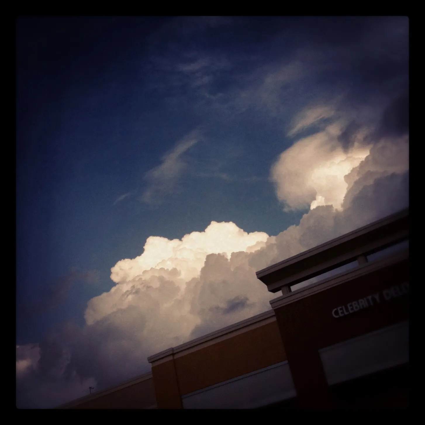 Dramatic view of large, billowing clouds at dusk above a shopping center building with the words “Celebrity Deli” partially visible.