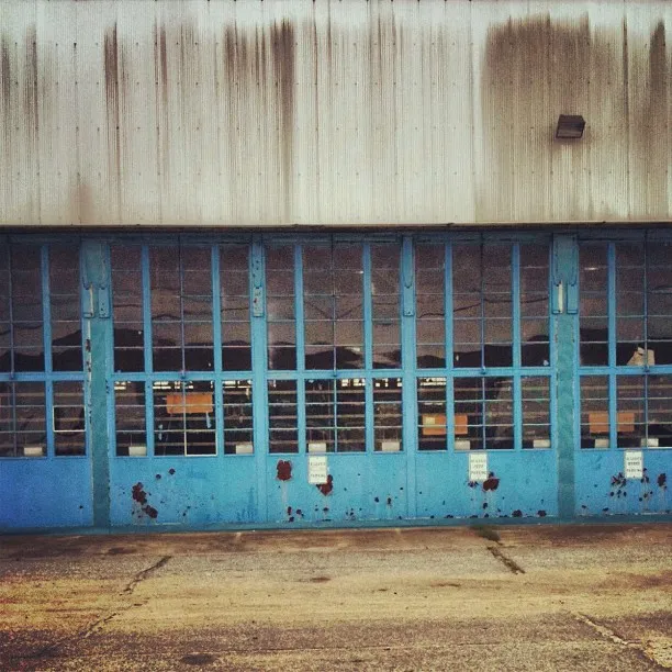 Front view of a weathered industrial building with large, blue, multi-paneled metal garage doors and streaked grime on the upper facade.