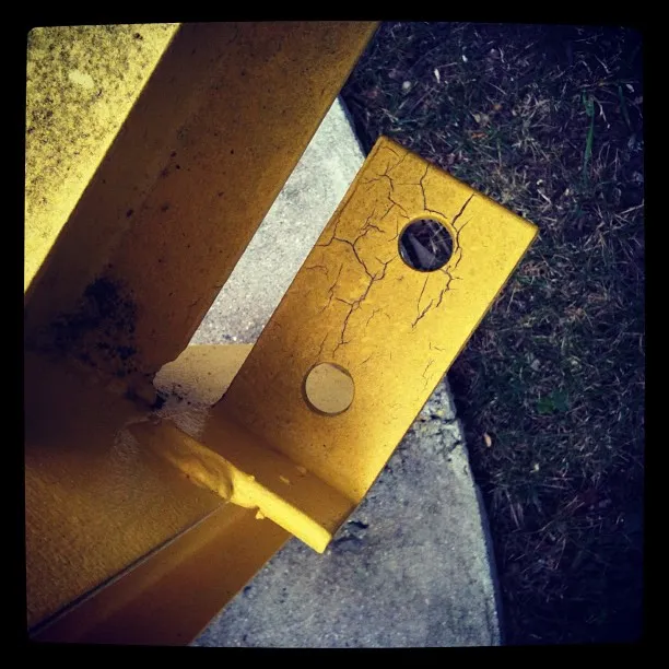 Close-up of a weathered yellow metal bracket with two holes, showing cracks in the paint near concrete and grass.