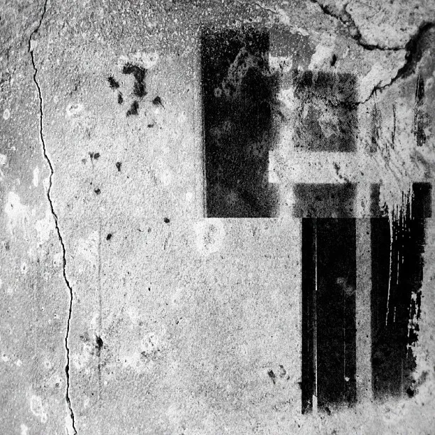 A cracked concrete surface with black rectangular and stencil-like spray paint marks, contrasting sharply against the textured gray background.
