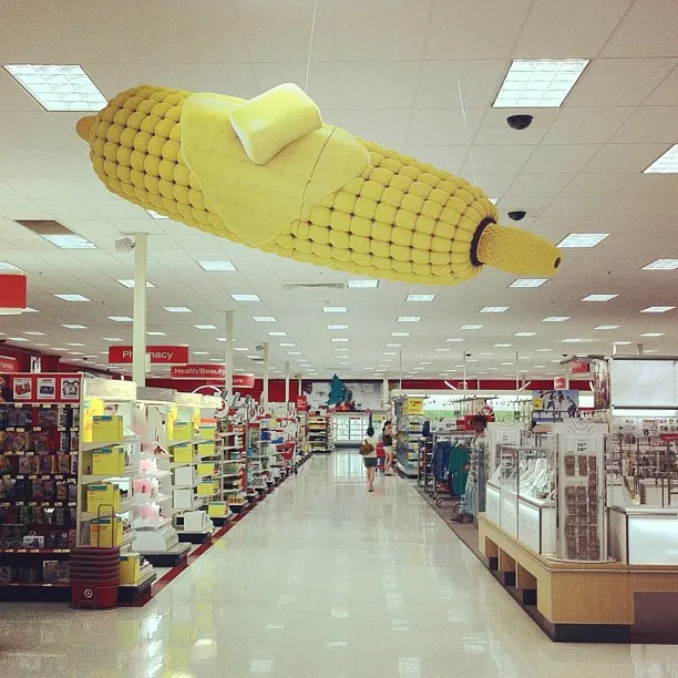 A giant model of a buttered corn on the cob is suspended from the ceiling inside a brightly lit retail store, above aisles of merchandise.
