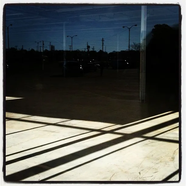 View through a reflective window showing a parking lot with streetlights and long interior shadows cast on a tiled floor.