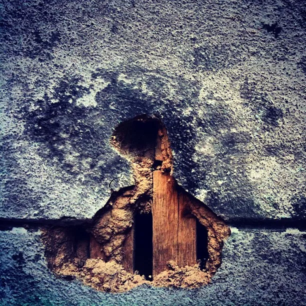 Crumbling plaster wall revealing wooden lath and dark cavity beneath, with hole shaped like a head and shoulders silhouette.