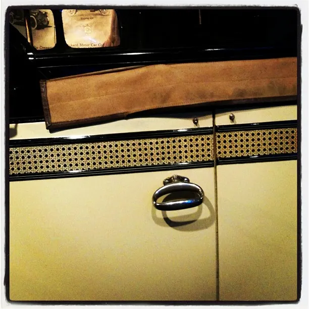 Close-up of a vintage car door with a woven pattern detail, chrome handle, and part of a tan convertible roof.