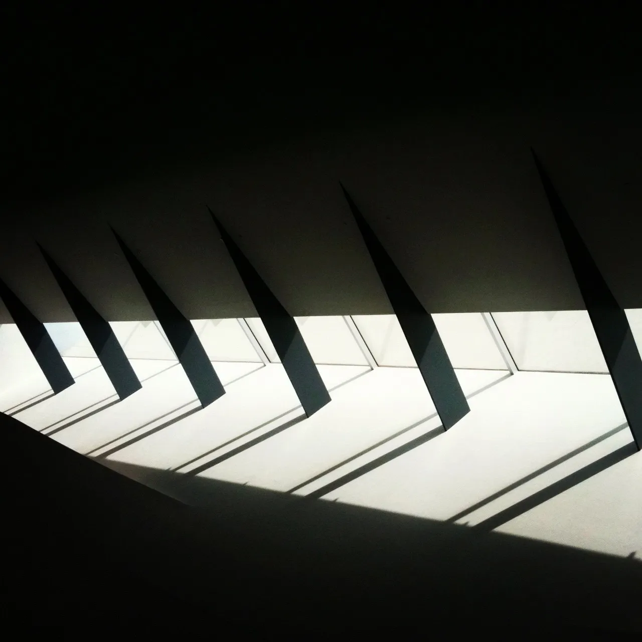 Interior ceiling with triangular cutouts casting angular shadows onto a white wall from overhead skylights.