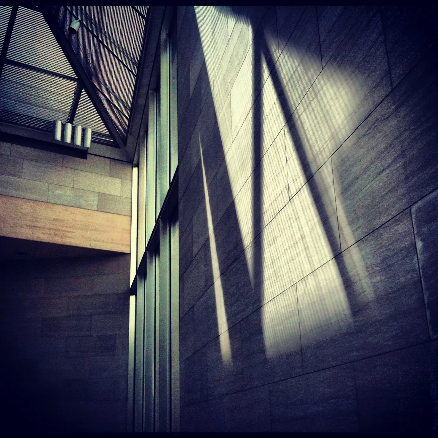 Interior view of a modern architectural space with tall windows and angled ceiling beams casting geometric shadows on a tiled wall.