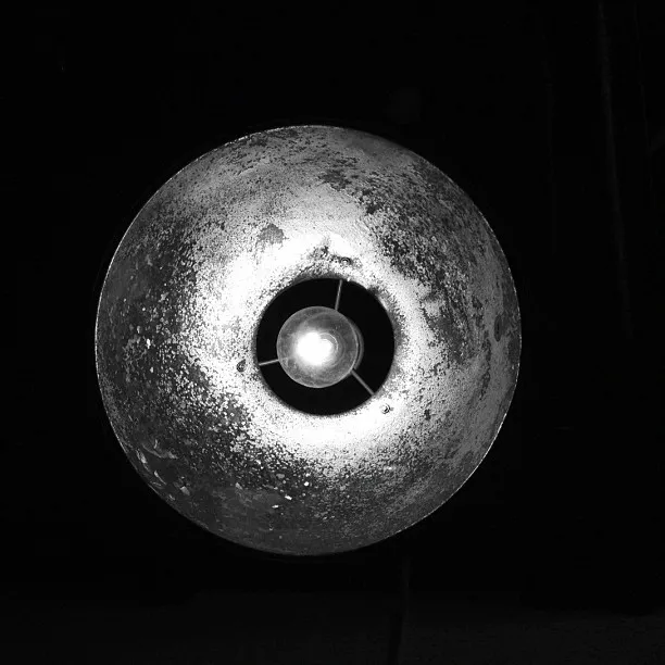 Close-up of a lit, weathered metal industrial lamp with a circular shade photographed in black and white against a dark background.