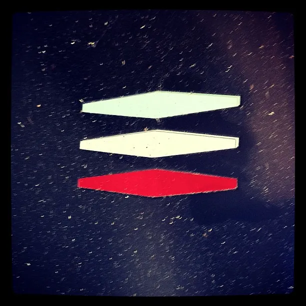 Three stacked rhombus shapes—two white and one red—on a speckled dark background, resembling a minimalist graphic symbol.